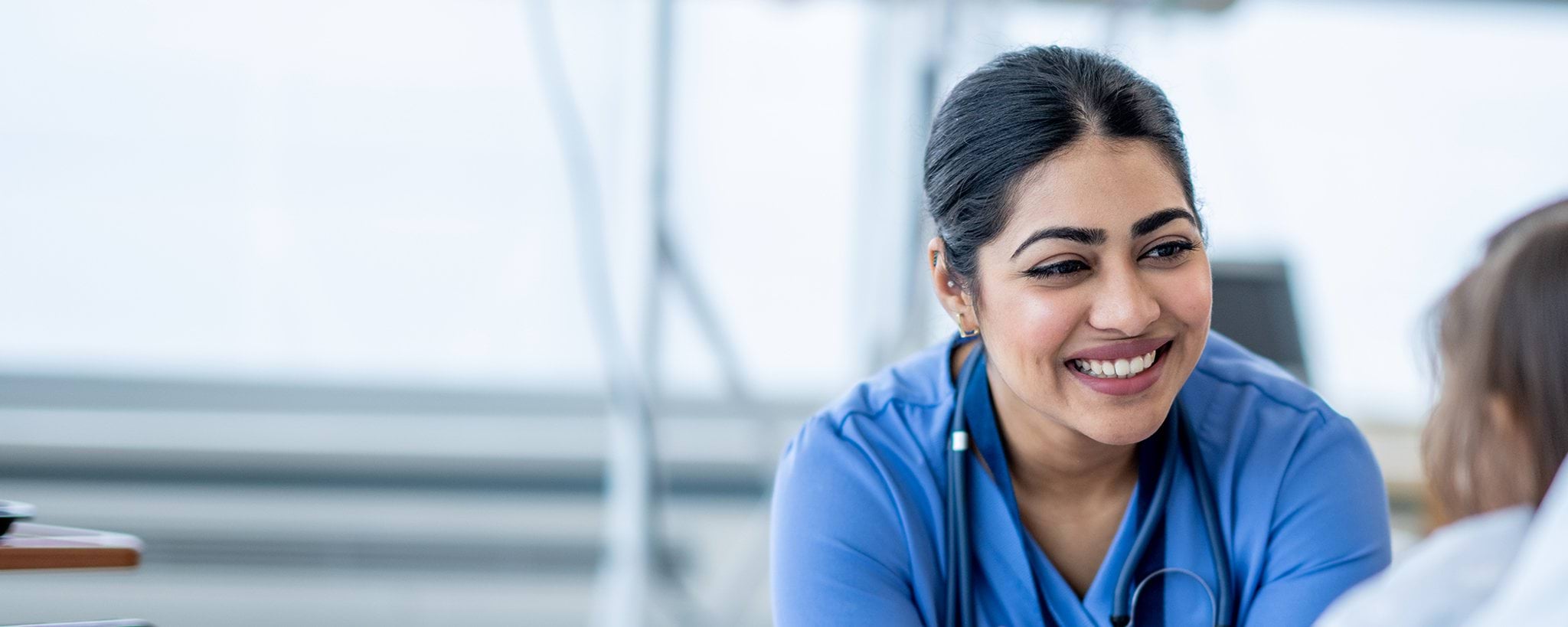A nurse in blue scrubs and a stethoscope smiles while interacting with a patient.