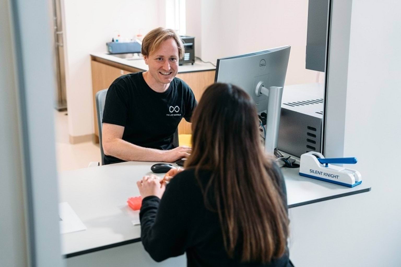 A man in a black t-shirt sits behind a desk and computer. He is talking to a woman with long brown hair.