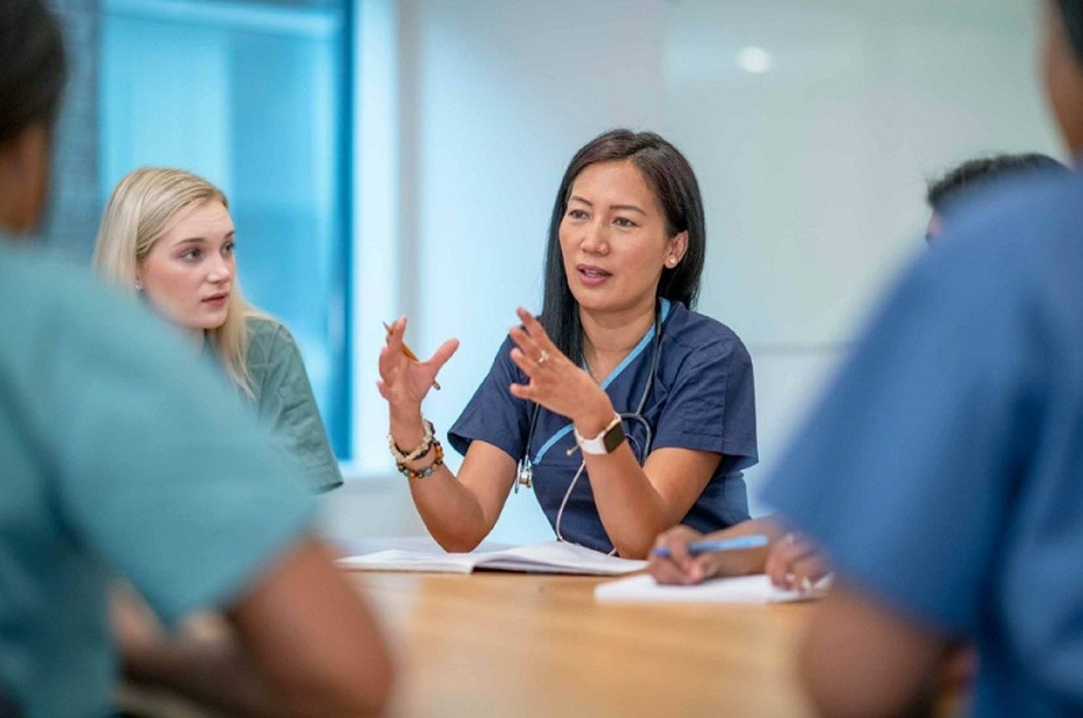 A woman wearing blue scrubs with a stethoscope. She is sitting at a table talking to her colleagues.