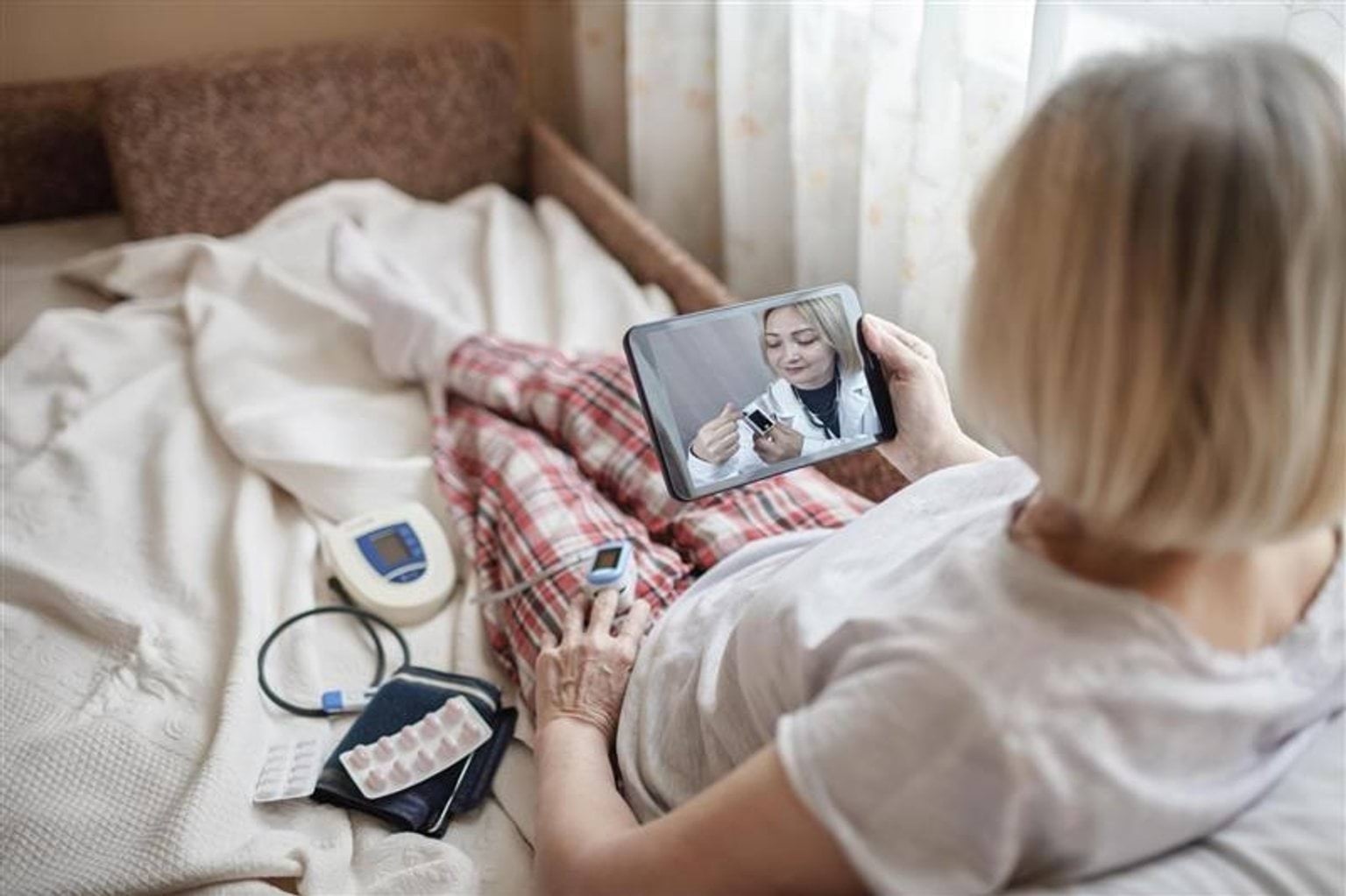 Person lying on a bed wearing plaid pyjama pants, holding a tablet showing a healthcare professional during a telehealth consultation as part of the Virtual Hospital pilot. A blood pressure monitor, a pulse oximeter clipped to one finger, and several blister packs of medication are placed nearby.