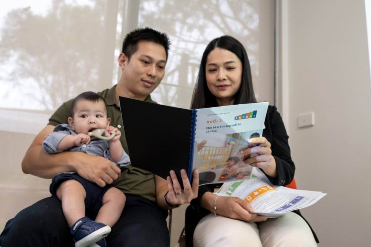A young family, with a mother, father and infant, reading a booklet together. The baby sits on the dads lap.