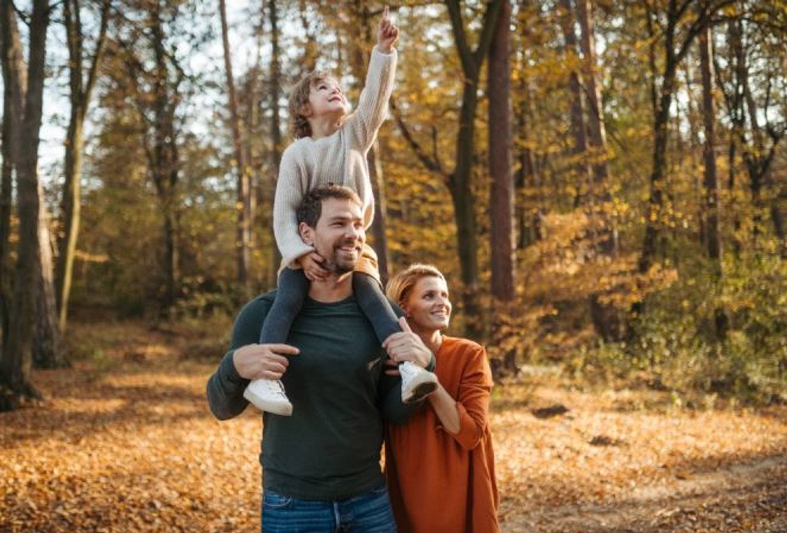 A family of a mother, father and young boy, walking in the woods during autumn, where the leaves surrounding them are orange.