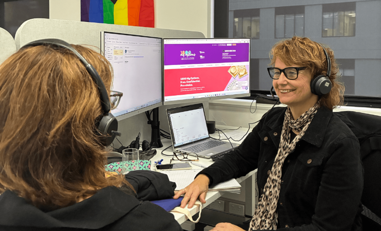 Two women at desks smile at each other