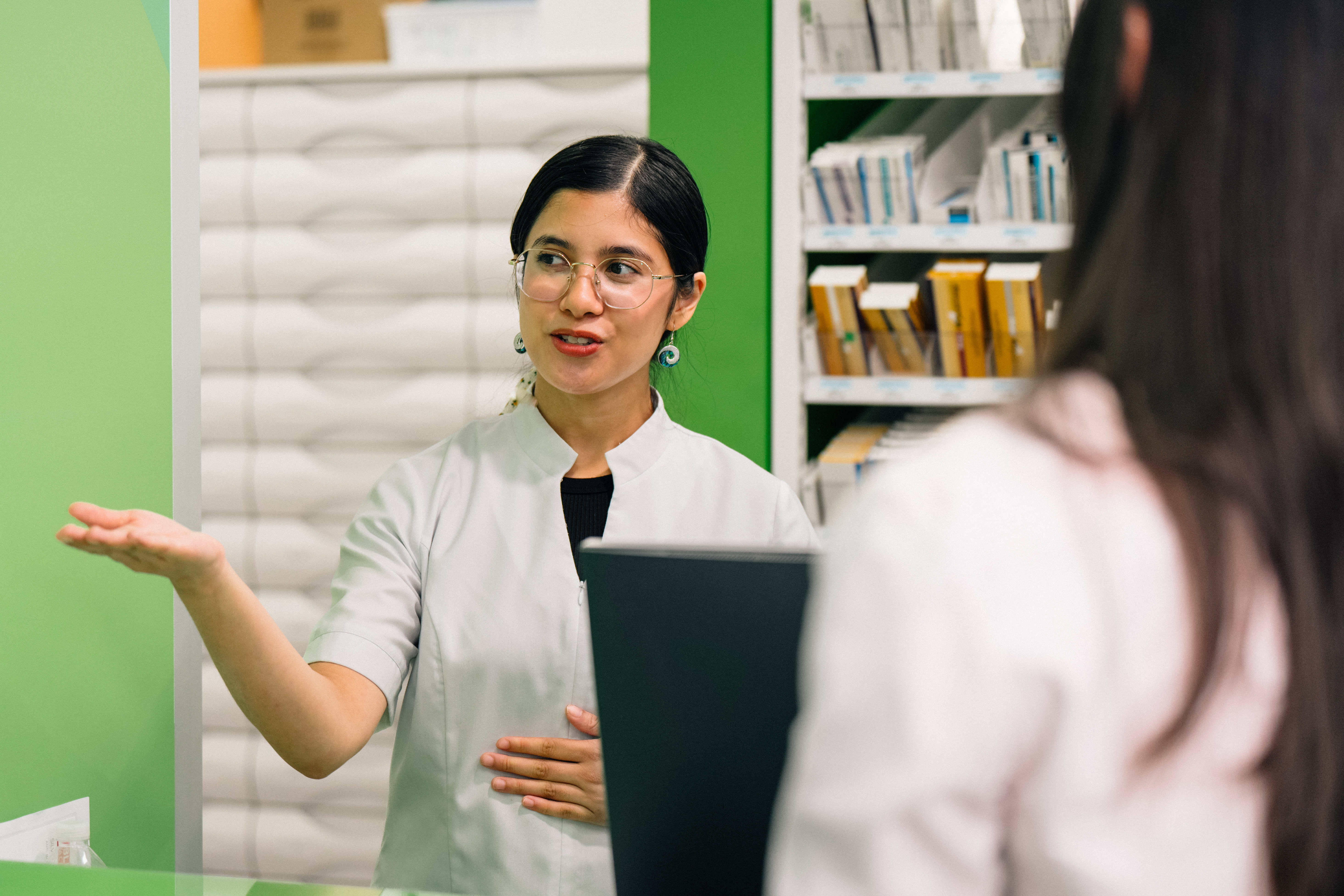 Person providing advice to a customer at a pharmacy counter, with medicine shelves visible in the background.