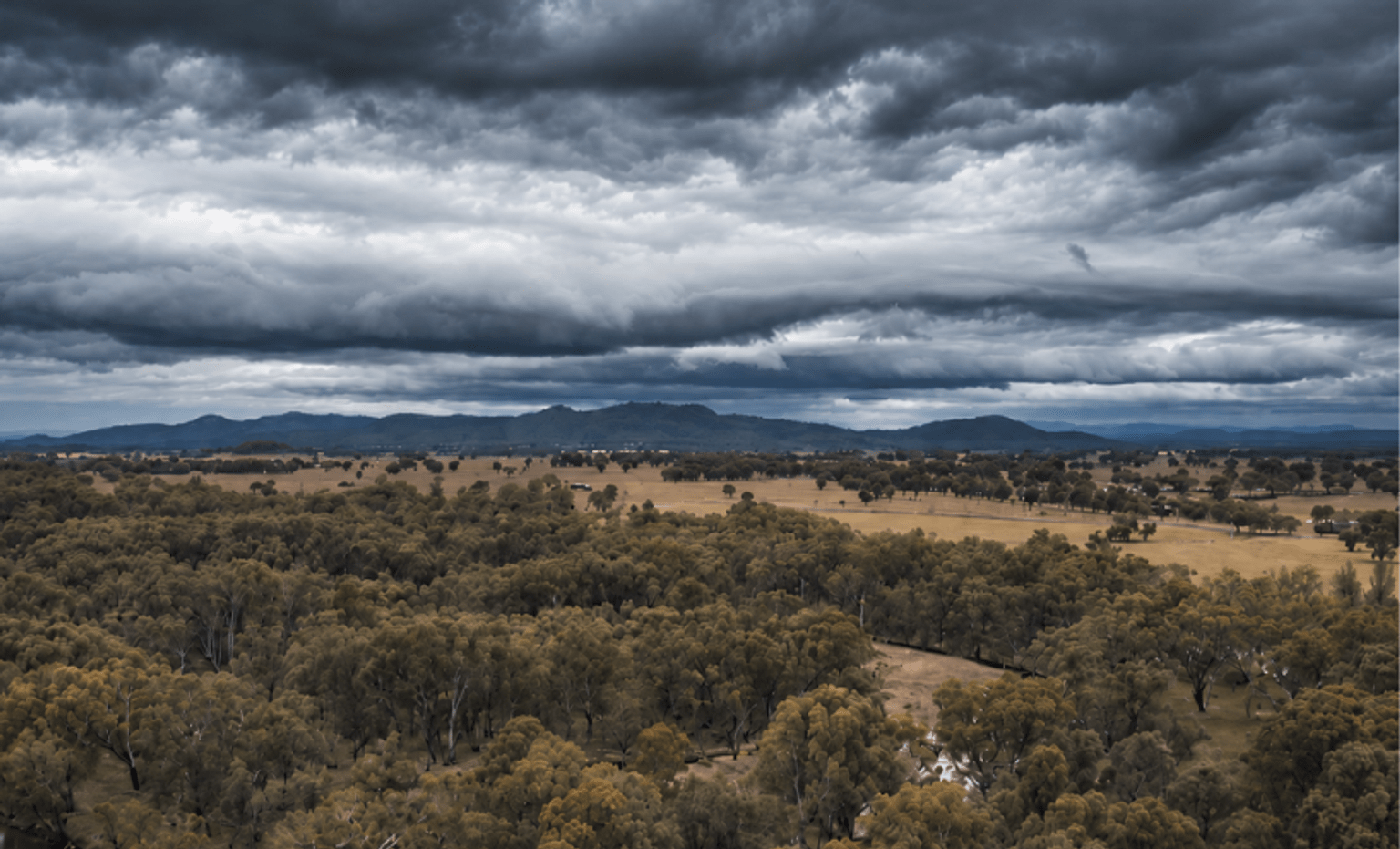 Storm clouds gathering over a rural landscape with farmland, trees and distant hills.